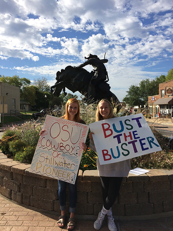 Two people hold signs in front of a large sculpture of a person on a bucking bronco. One sign says "OSU cowboy? Or Stillwater Pioneer?" The other sign says "bust the buster."